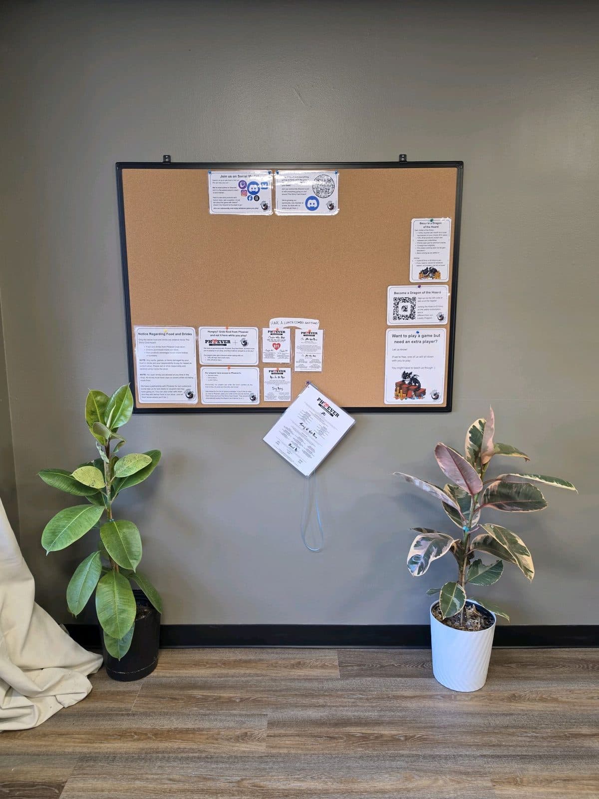 A cork noticeboard between two potted plants, pinned with event flyers, a Discord invite and a takeaway menu.